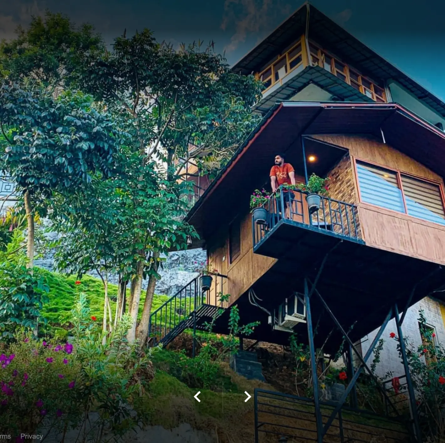 Stilted wooden hut at dusk with a guest standing on the balcony above the hillside